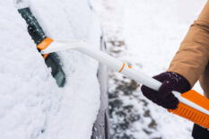 Woman cleans snow from a car with a scraper and a brush